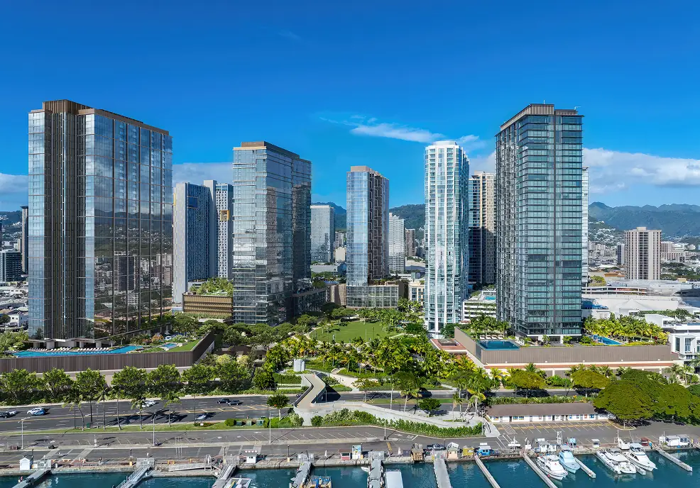A cluster of modern high-rise buildings with glass exteriors stands near a marina, with mountains and a blue sky in the background.
