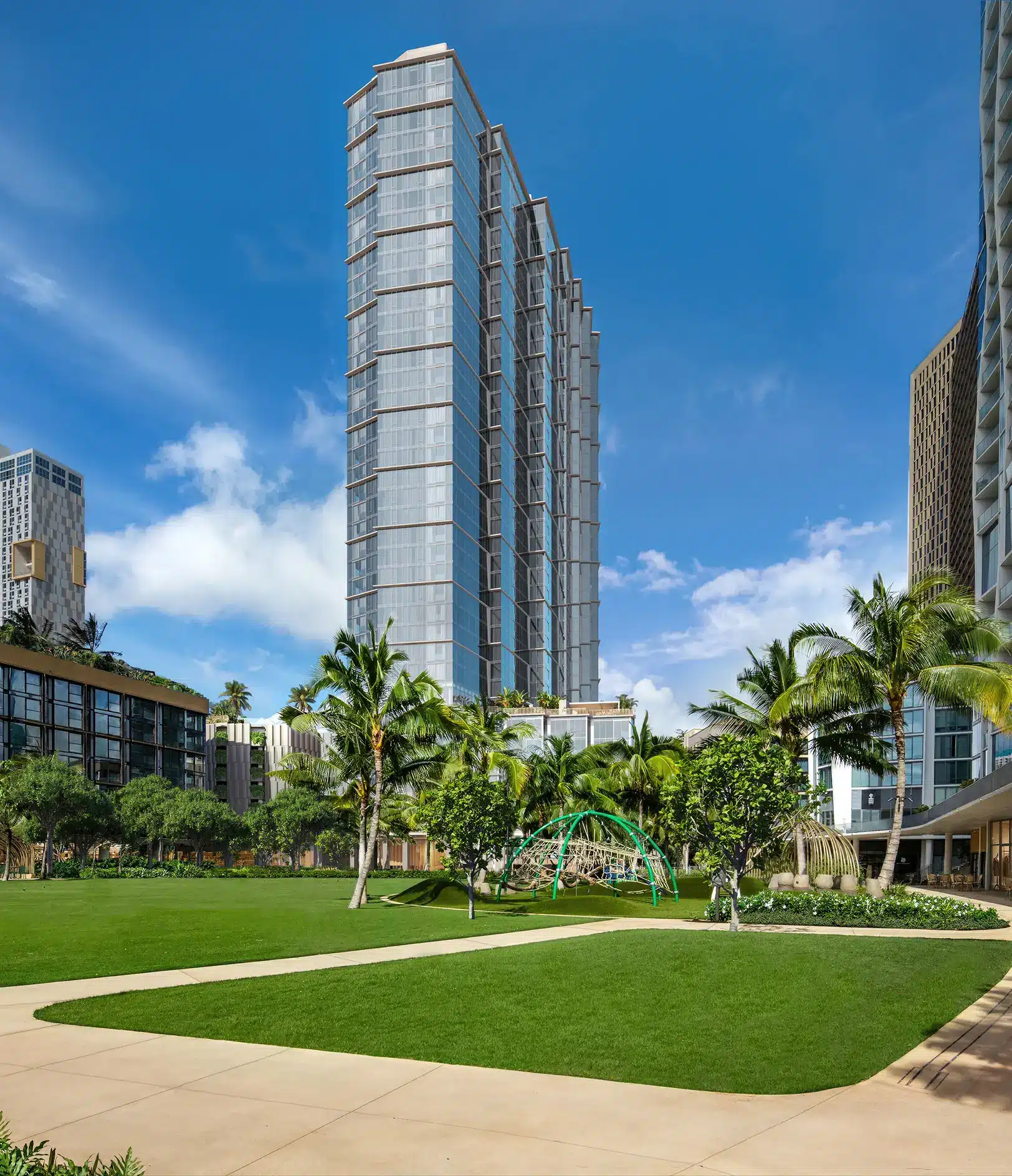 Tall modern glass building surrounded by palm trees, green lawn, and pathways under a blue sky with scattered clouds.