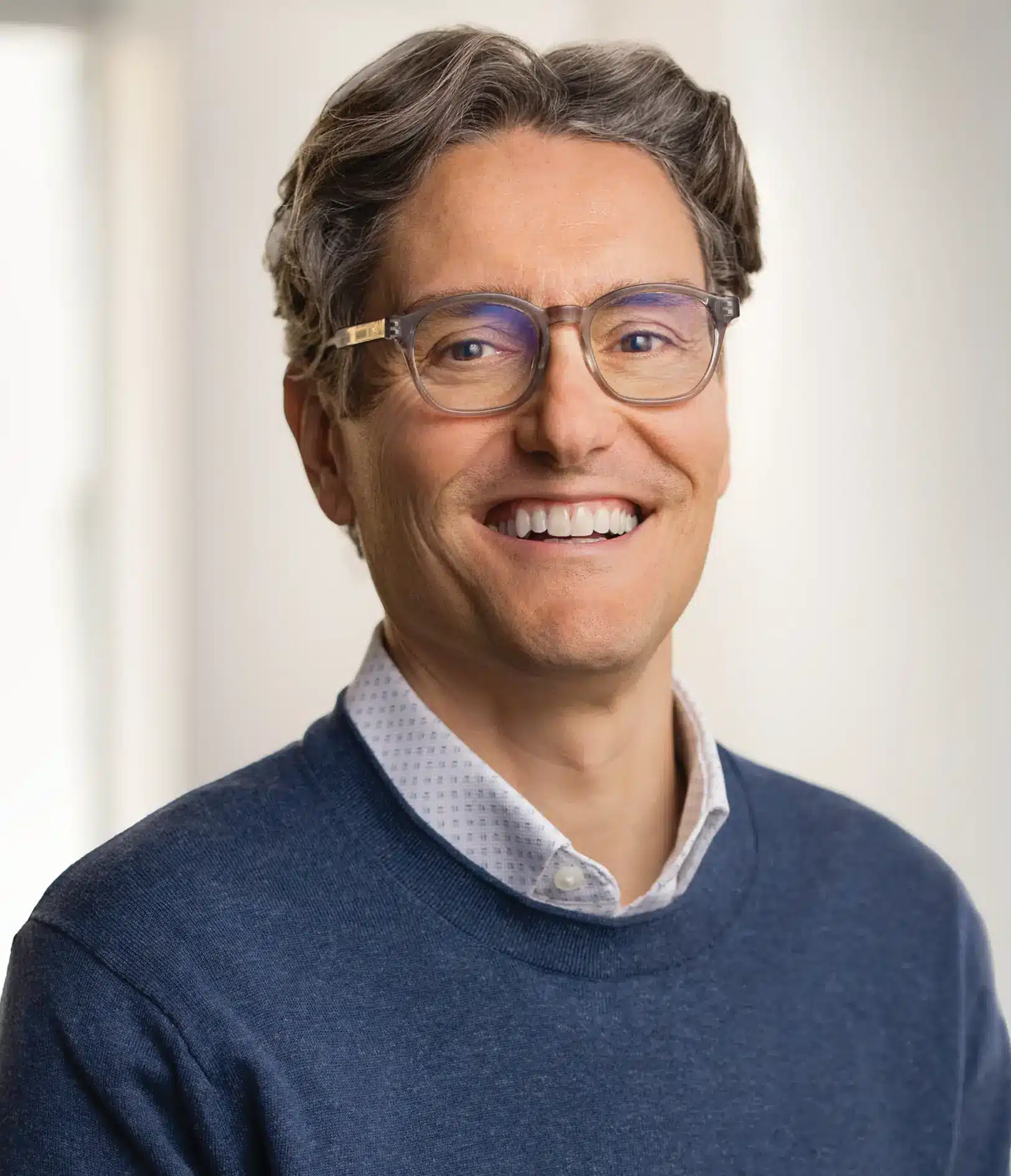 Smiling man with wavy brown hair and glasses wearing a blue sweater over a collared shirt, standing in a bright indoor setting.