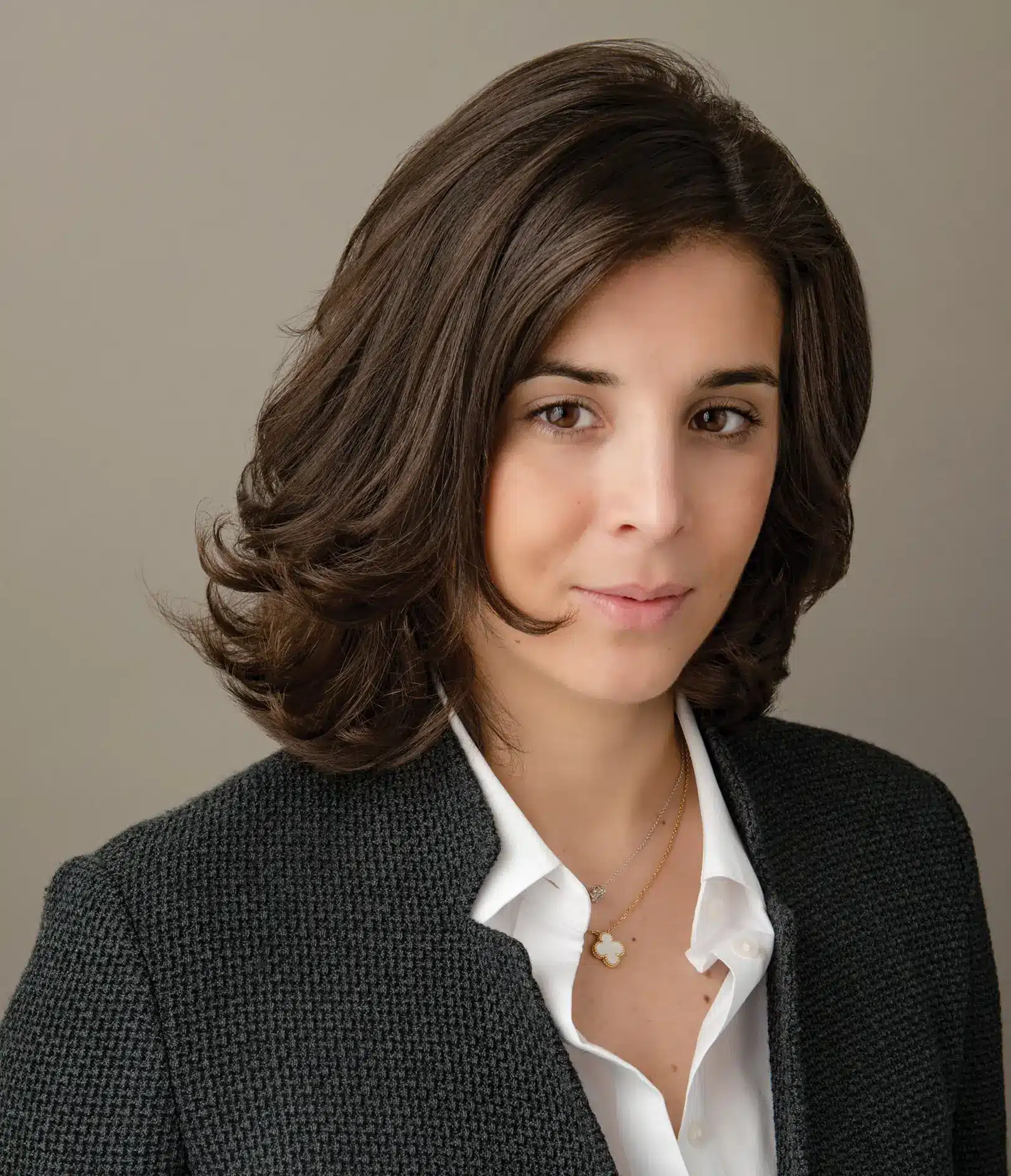 Woman with shoulder-length brown hair wears a dark textured blazer over a white blouse, standing against a plain neutral background and looking at the camera.