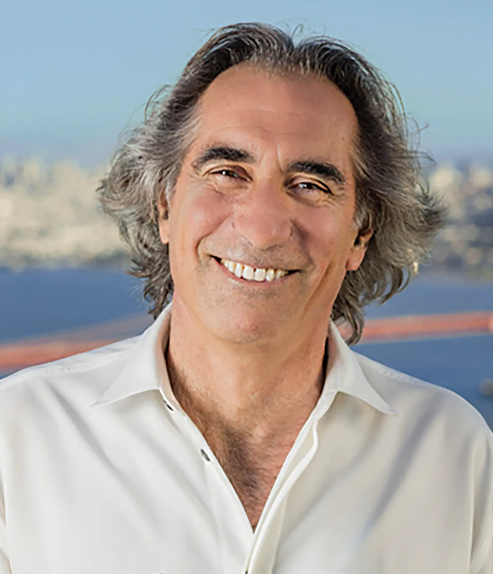 A middle-aged man with gray wavy hair wearing a white shirt, smiling in front of a blurred cityscape and bridge.