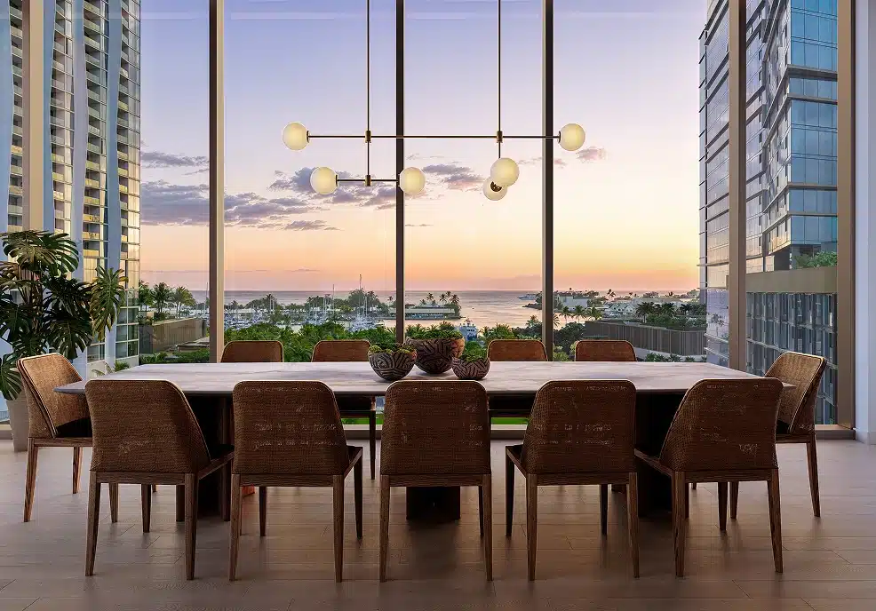Modern dining room with a long table and ten chairs, large glass windows, contemporary chandelier, and a sunset view of a marina and palm trees outside.