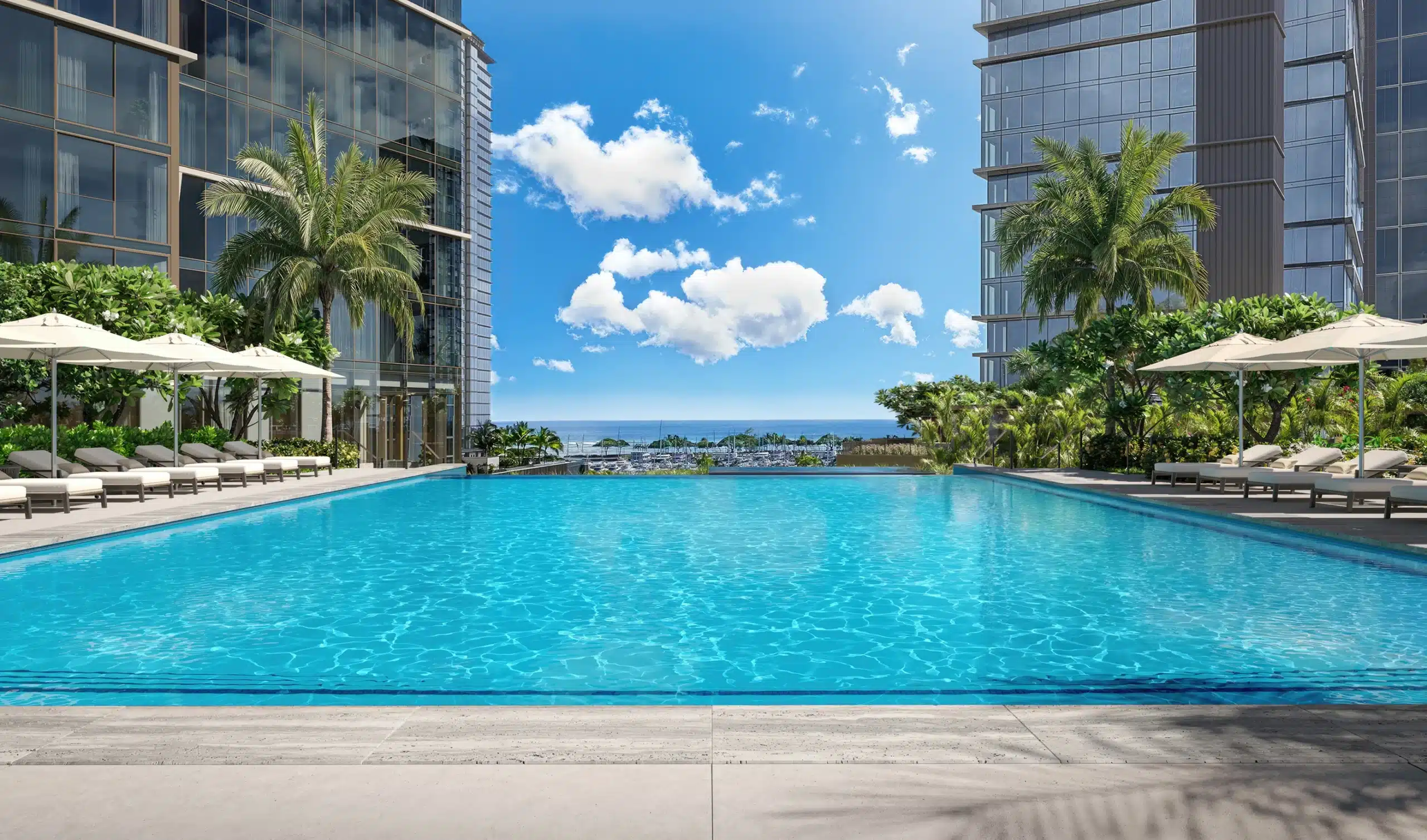 Rectangular outdoor swimming pool with lounge chairs, palm trees, and umbrellas, situated between two modern glass buildings, overlooking the ocean under a blue sky with scattered clouds.