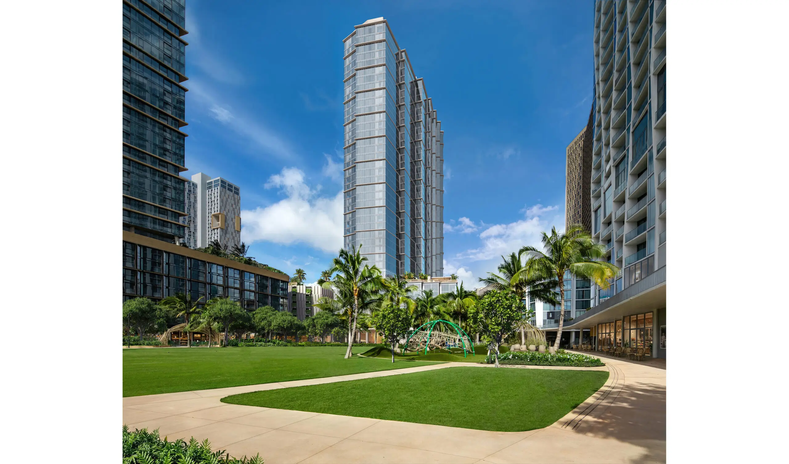 Modern high-rise buildings surround a landscaped courtyard with green grass, palm trees, walking paths, and a small playground under a blue sky with scattered clouds.
