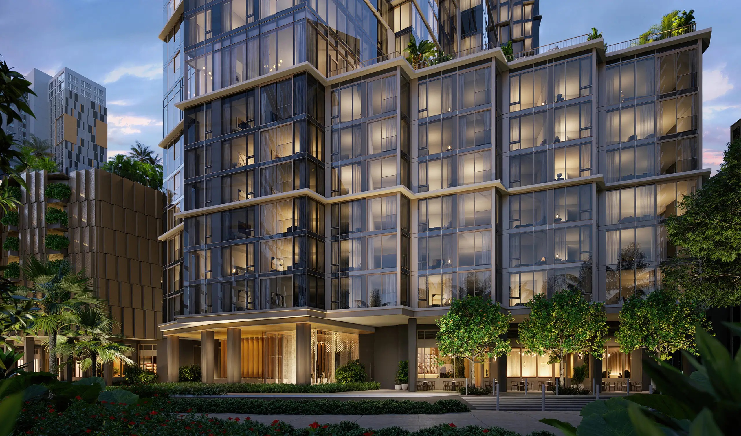 Modern multi-story building with large glass windows, exterior lighting, and landscaped greenery in the foreground, photographed at dusk.
