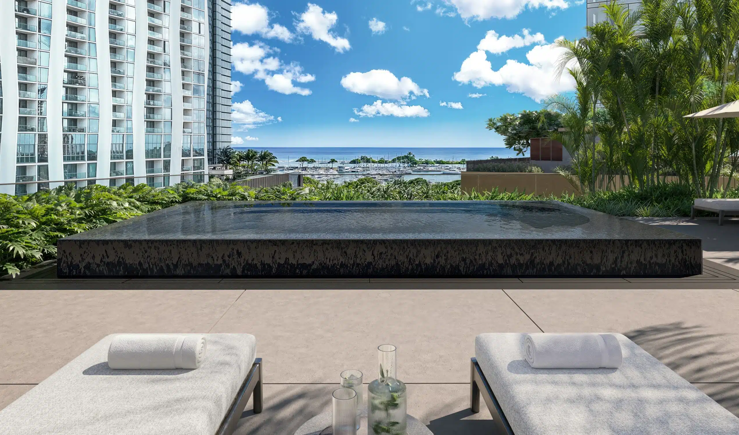 Infinity pool on a rooftop deck with two lounge chairs in the foreground, overlooking a cityscape, greenery, and distant ocean under a blue sky with clouds.