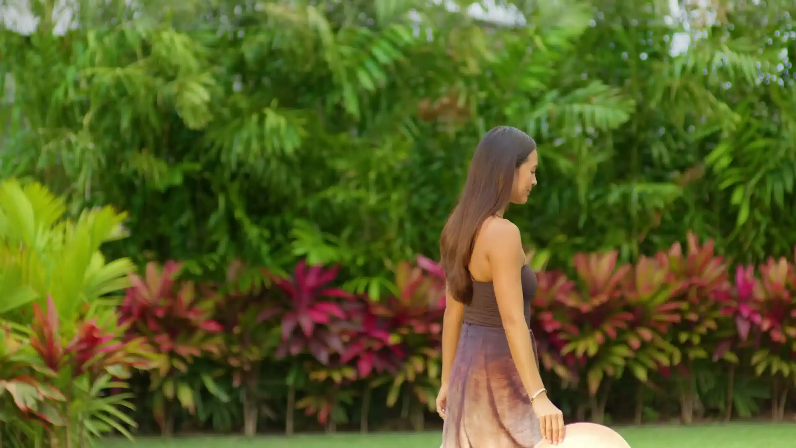 A woman in a brown dress carries a hat while walking outdoors in front of lush green and colorful tropical plants.