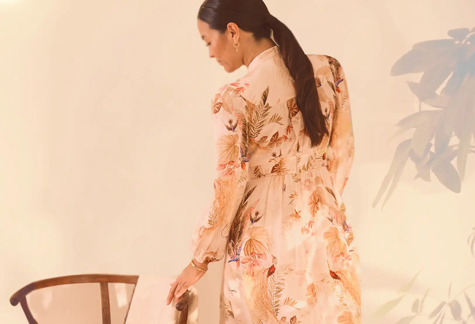 Woman in a light-colored floral dress stands beside a wooden chair, viewed from behind, with her hair in a low ponytail against a neutral background.