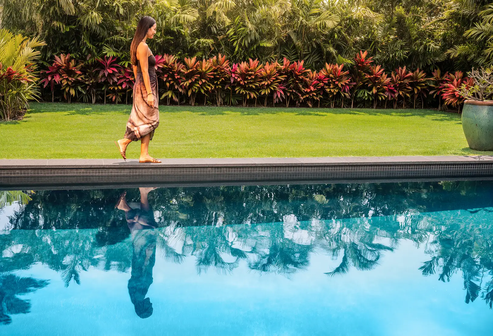 A woman in a long dress walks beside a swimming pool with her reflection visible in the water; lush plants and grass are in the background.