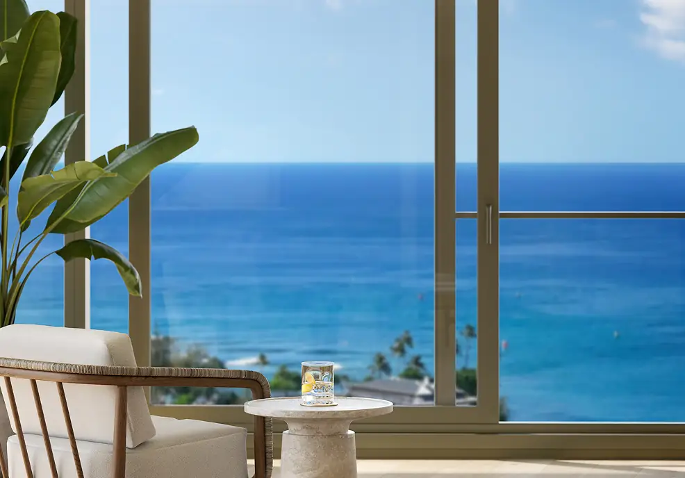 A glass of water with lemon sits on a small table near a chair and a large window overlooking the ocean on a clear day.