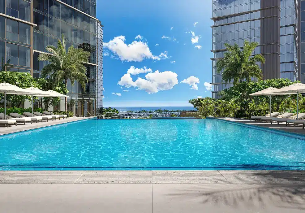 Large outdoor pool between two modern glass buildings, surrounded by lounge chairs, umbrellas, palm trees, and with an ocean view in the background under a blue sky with clouds.