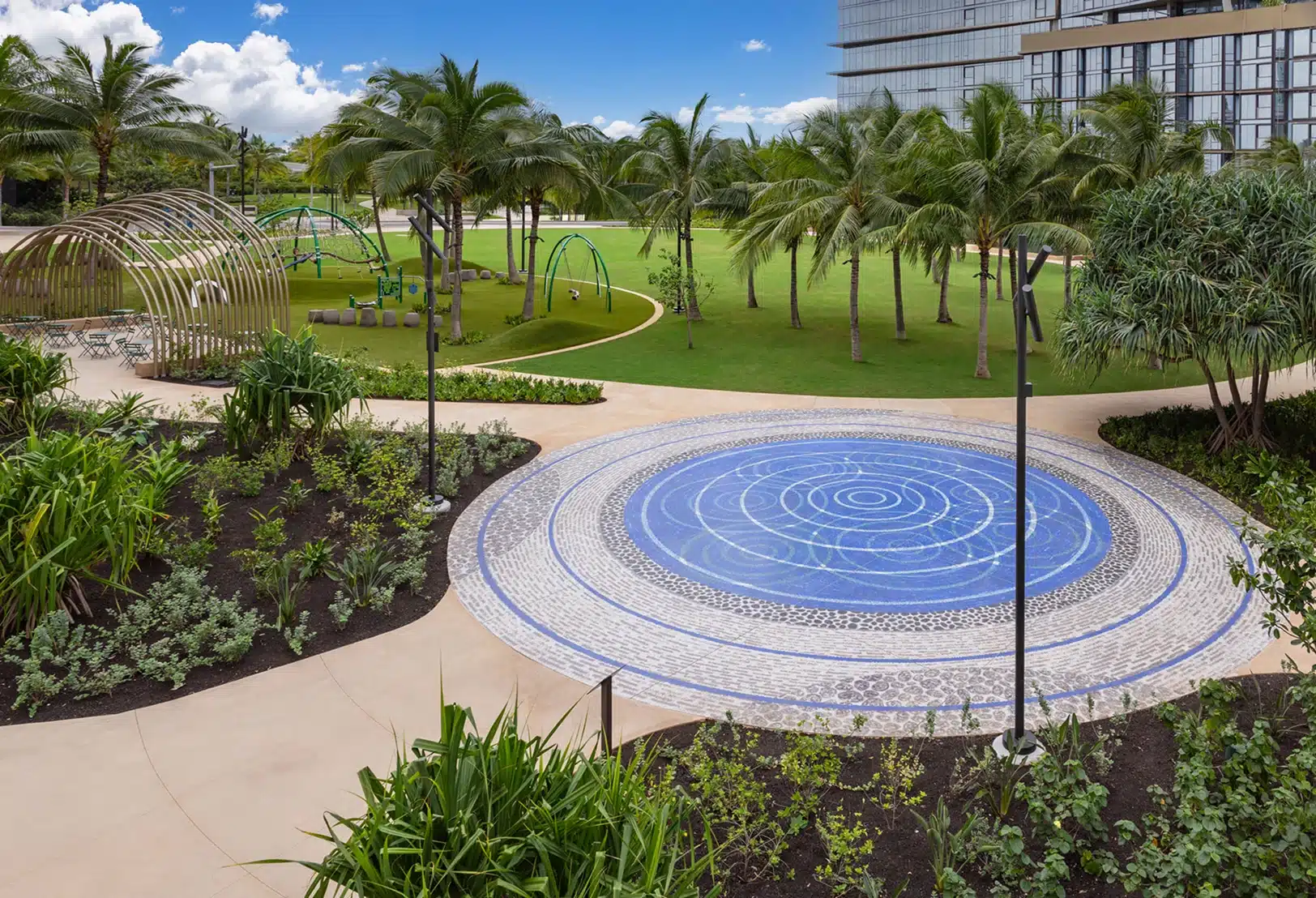Circular blue mosaic water feature on a landscaped path, surrounded by green grass, palm trees, modern playground structures, and a glass building in the background under a blue sky.