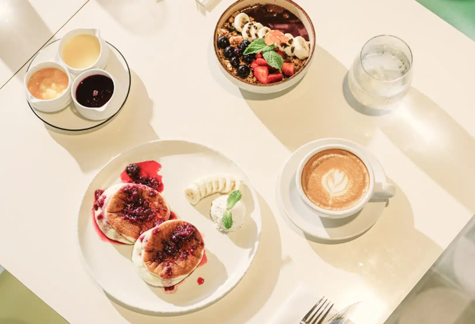 A breakfast table with pancakes, berry sauce, banana, whipped cream, smoothie bowl with fruit, a cup of latte with latte art, two small sauces, a fork, knife, and a glass of water.
