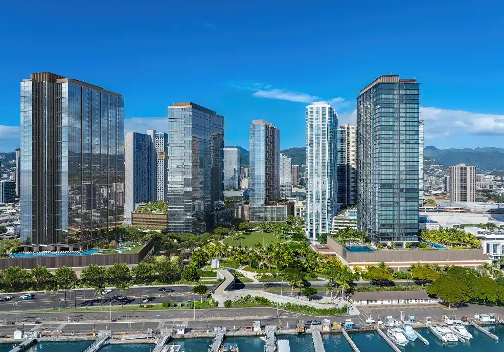 A group of modern high-rise buildings stands near a marina under a clear blue sky, with mountains visible in the distant background.