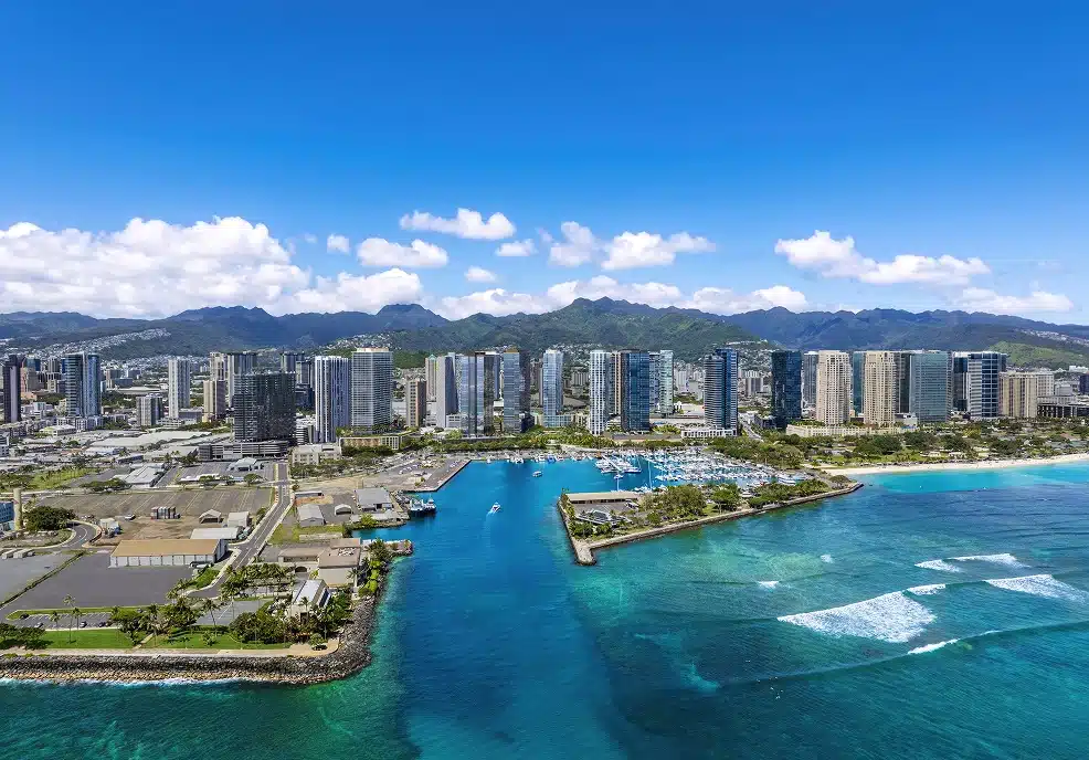 Aerial view of a coastal city skyline with high-rise buildings, clear blue water, small waves, and mountains in the background under a sunny sky.