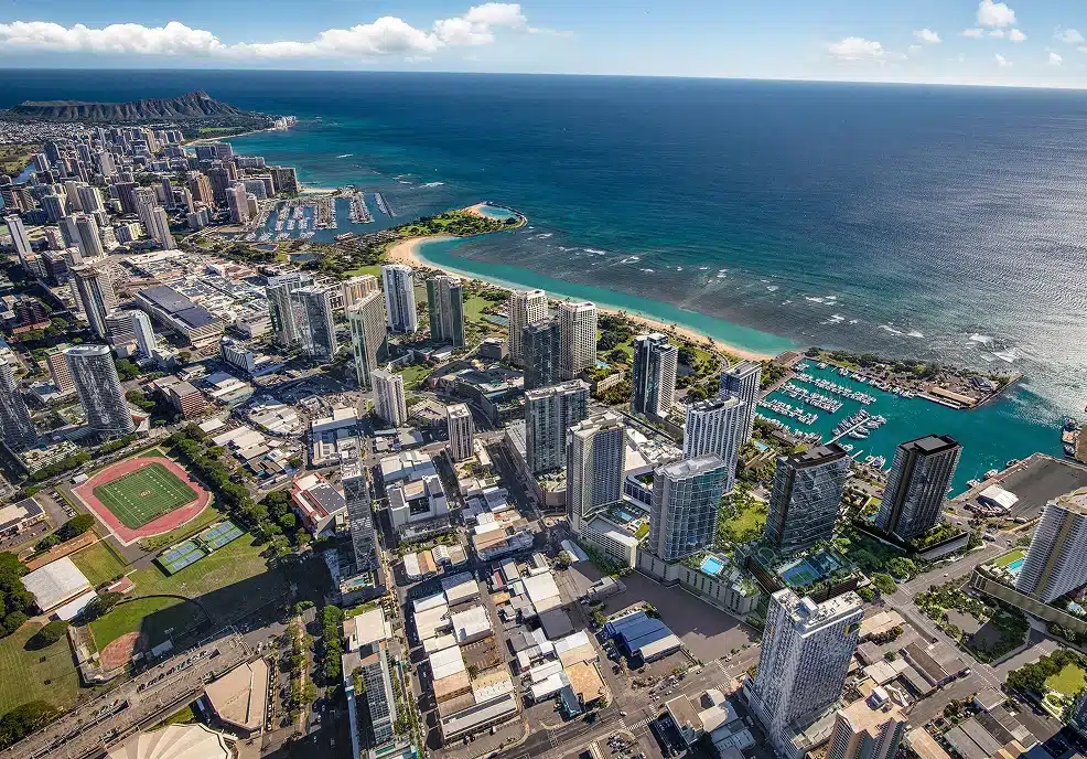 Aerial view of a coastal city with high-rise buildings, a sandy beach, a marina, and mountains in the background under a partly cloudy sky.