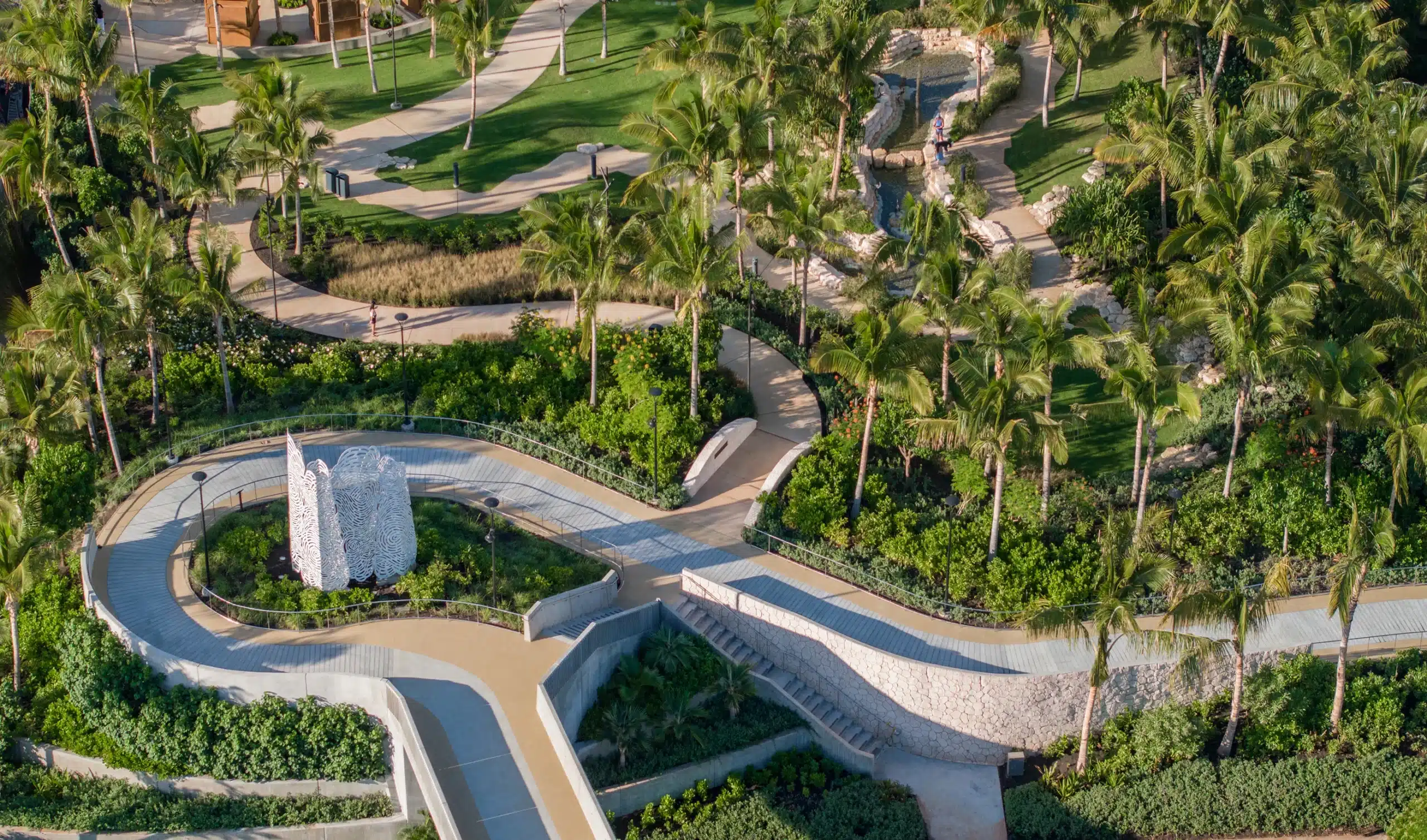 Aerial view of a landscaped park with winding paths, palm trees, green lawns, and a white stone sculpture surrounded by tropical plants.