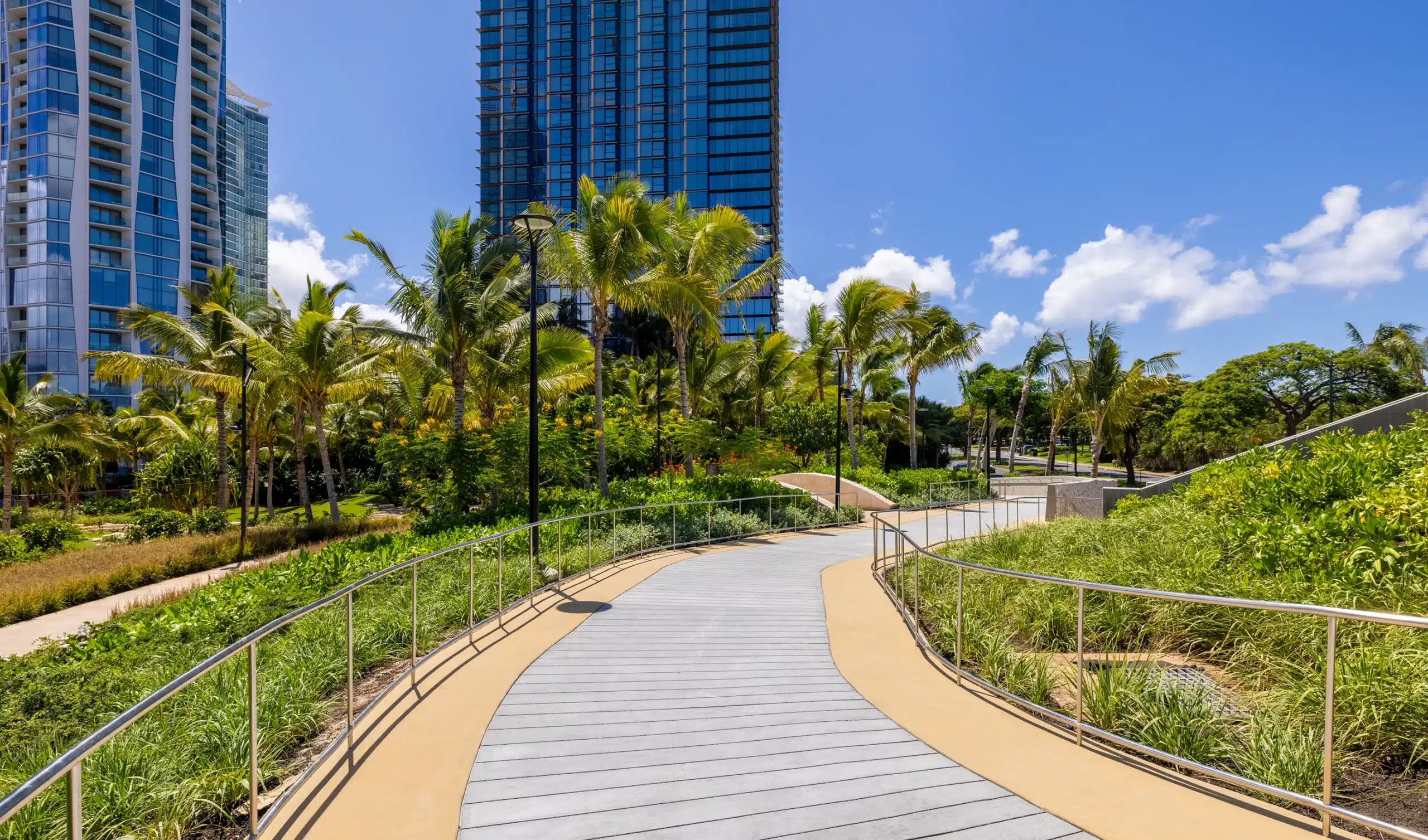 Curved pathway with railings winds through landscaped greenery, palm trees, and tall modern buildings under a clear blue sky.