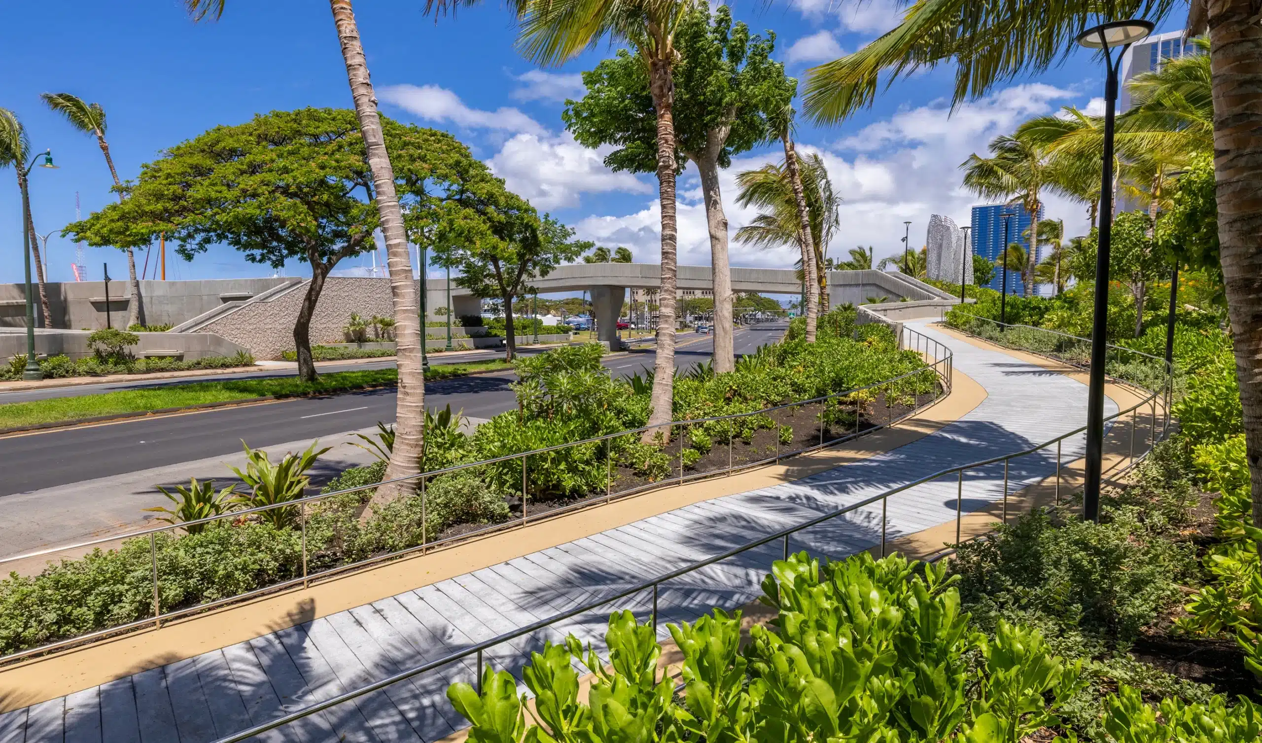 Curved pedestrian walkway with railings beside a road, surrounded by palm trees, lush greenery, and blue sky with a few clouds.