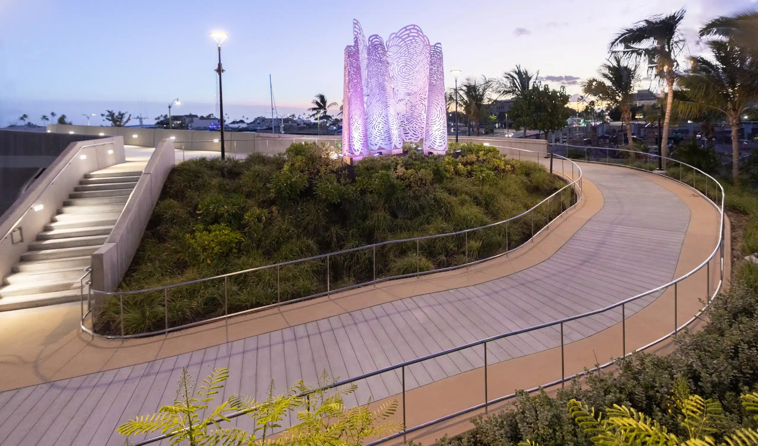 Curved walkway with metal railings surrounds a grassy mound featuring a lit purple sculpture, taken at dusk with palm trees in the background.