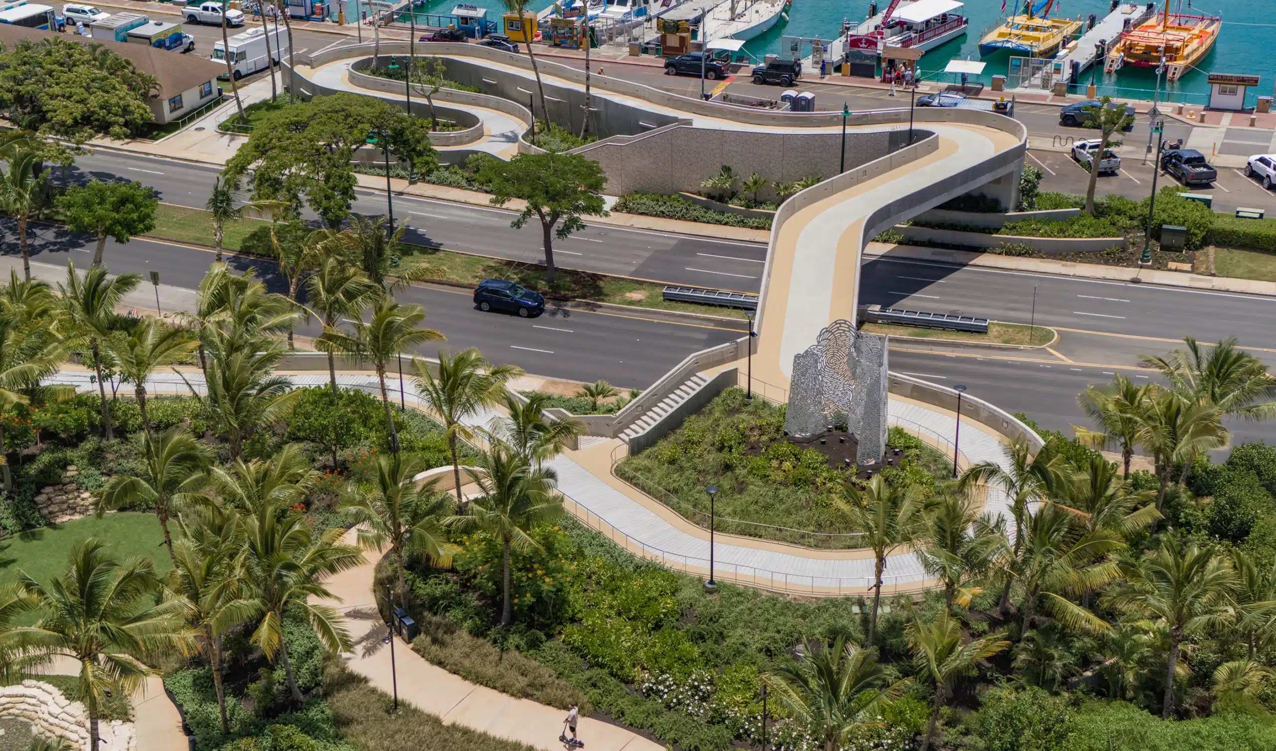 Curved pedestrian bridge crosses over a busy road, surrounded by greenery, palm trees, and a nearby marina with boats.
