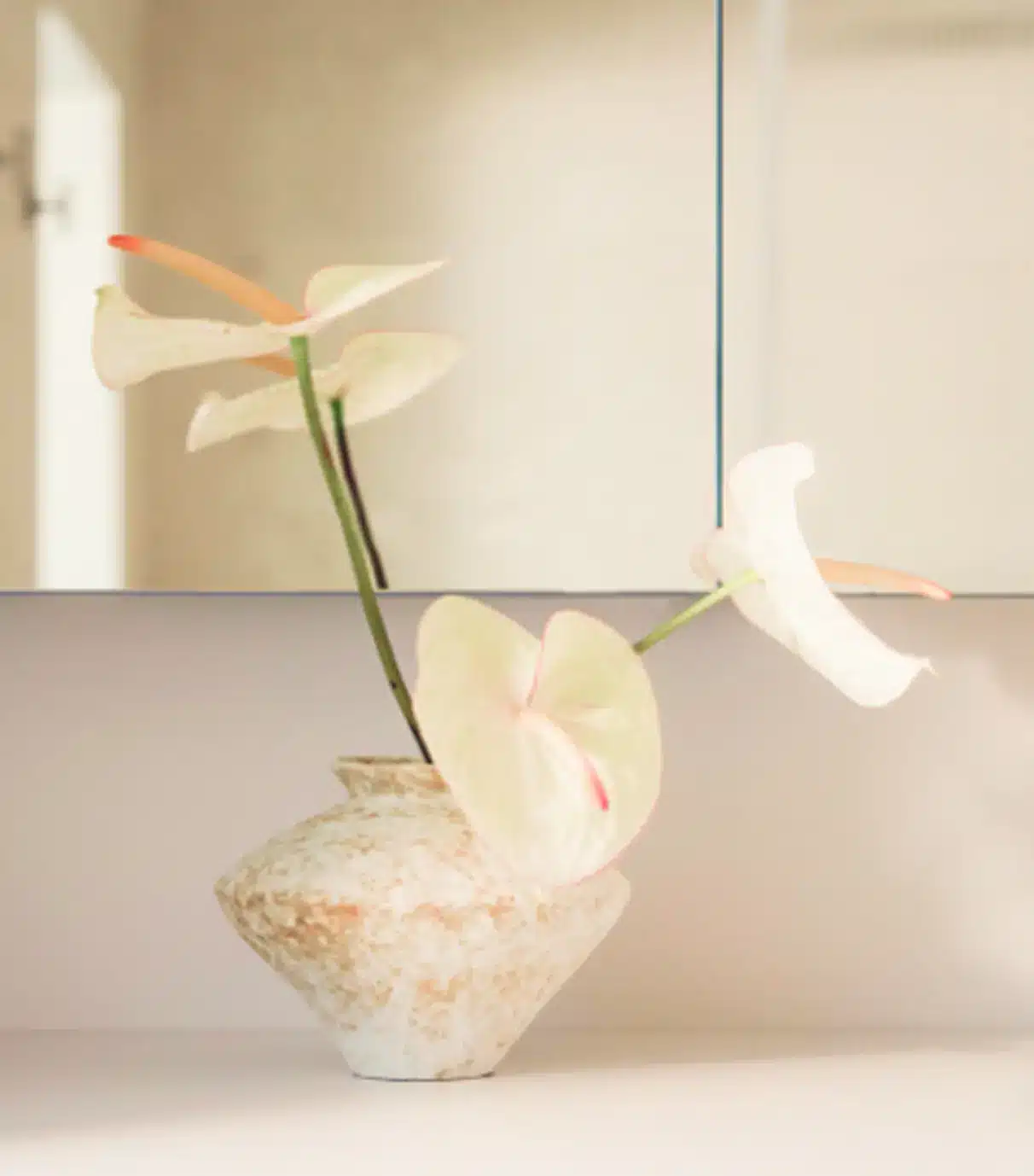 A ceramic vase with three white anthurium flowers sits on a light-colored surface in front of a mirror.