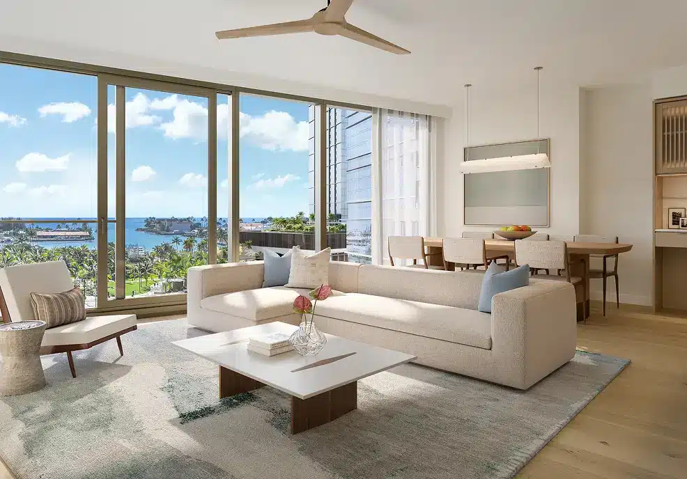 Modern living room with large windows, beige sofa, coffee table, dining area, and a view of a harbor and palm trees outside on a sunny day.