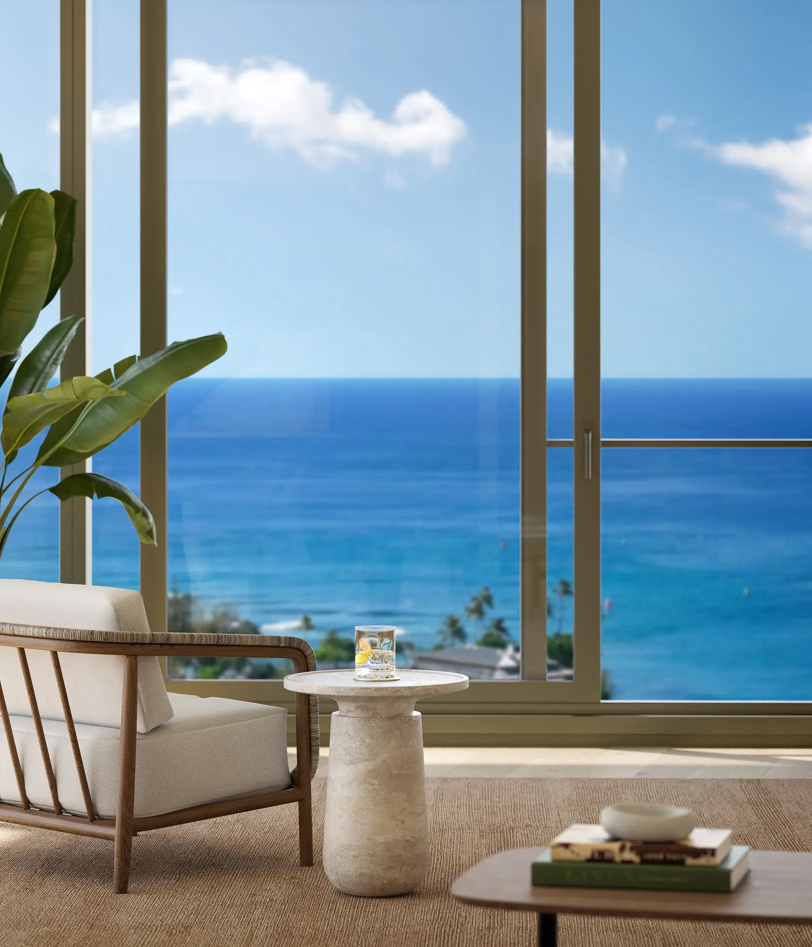 A lounge chair and small table sit by large windows overlooking a blue ocean and sky, with a plant and books nearby.