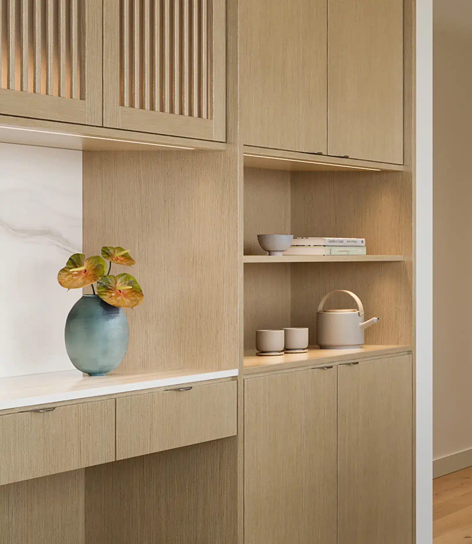 Light wood cabinets with open shelves display a blue vase with leafy plant, a teapot, cups, and stacked books; white marble accent is visible in the background.