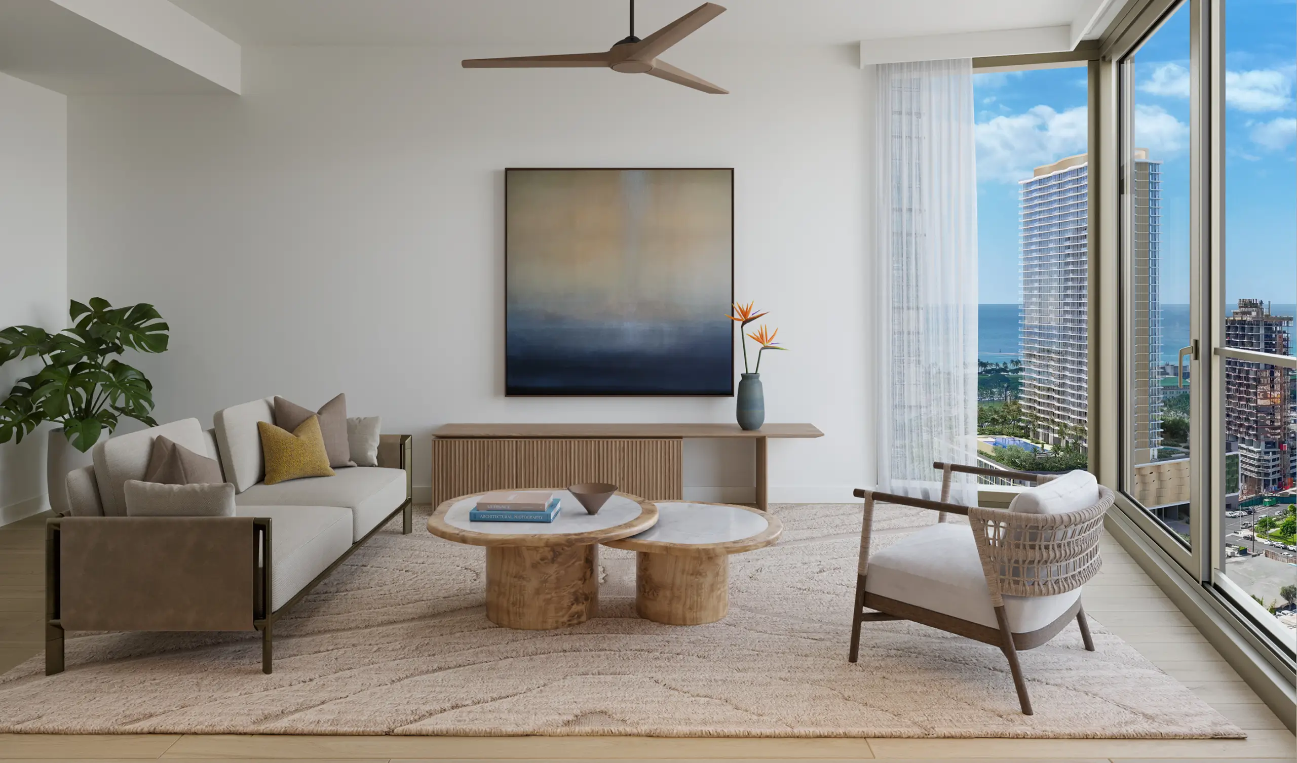 Modern living room with a sofa, armchair, coffee tables, abstract wall art, large window with city and ocean view, and a ceiling fan. Neutral tones and natural light dominate the space.