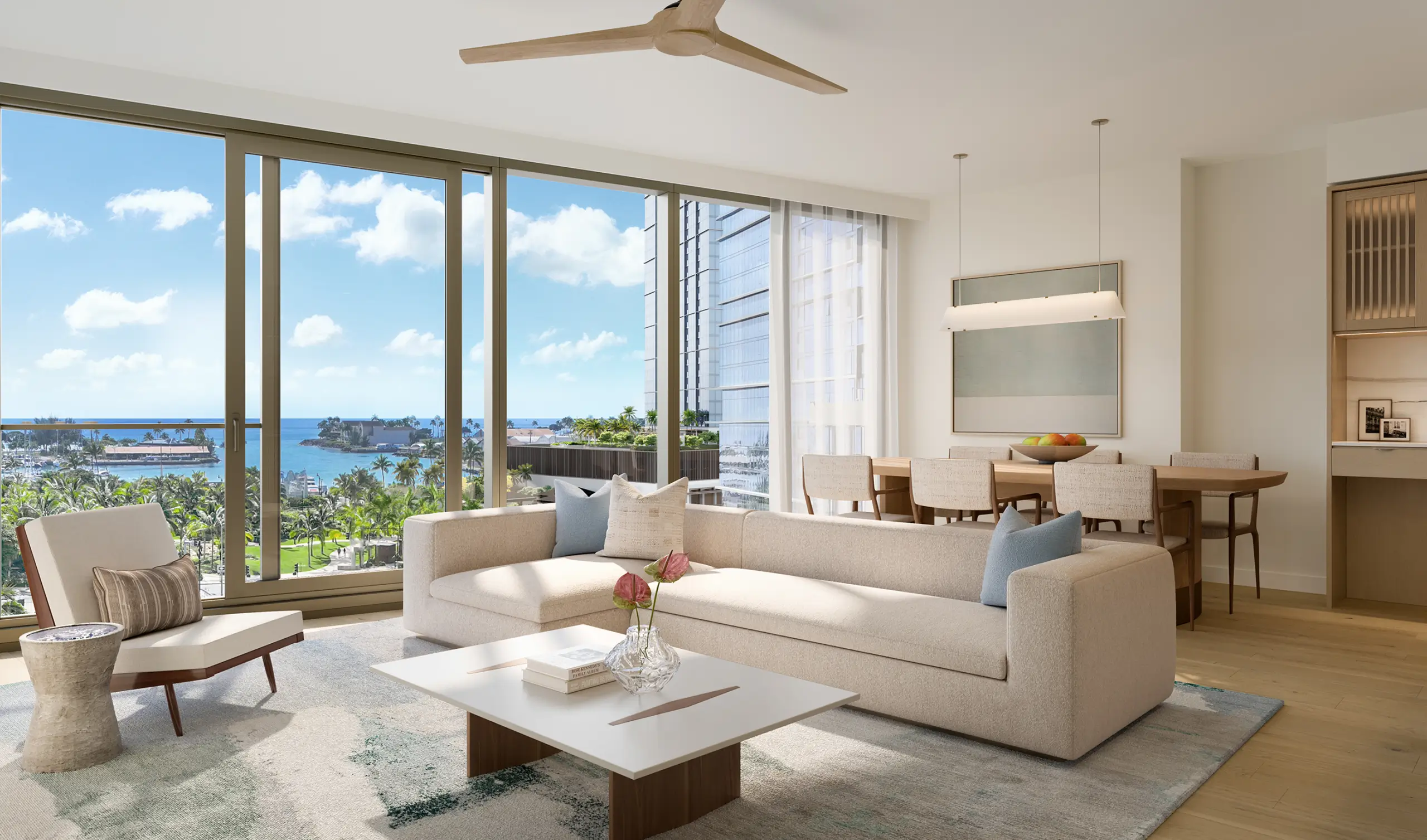 Modern living room with beige sofa, coffee table, large windows showing ocean view, and dining area with table and chairs in the background.