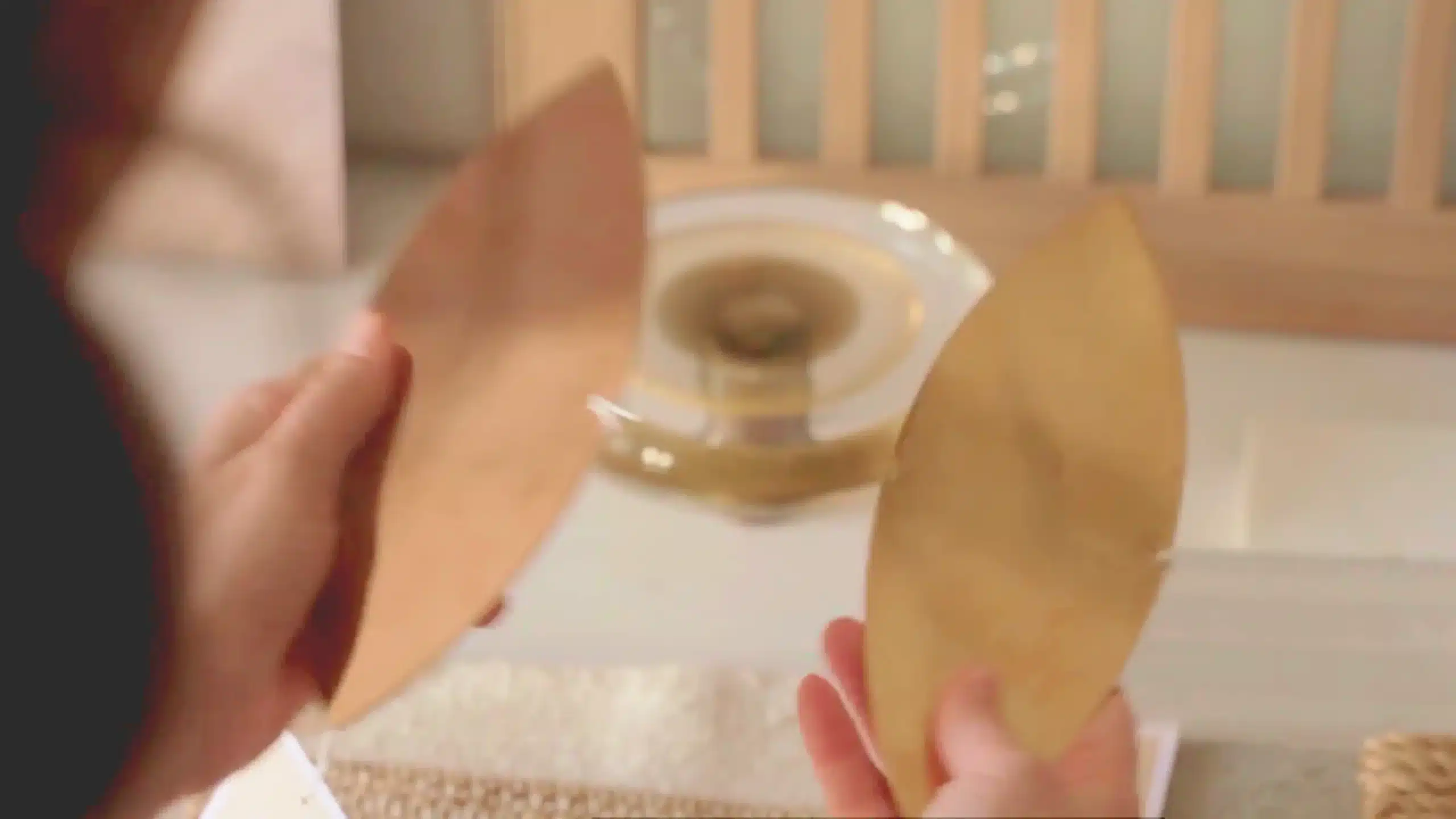 Two hands hold gold-colored, leaf-shaped objects in front of a blurred background featuring a circular glass object and wooden slats.