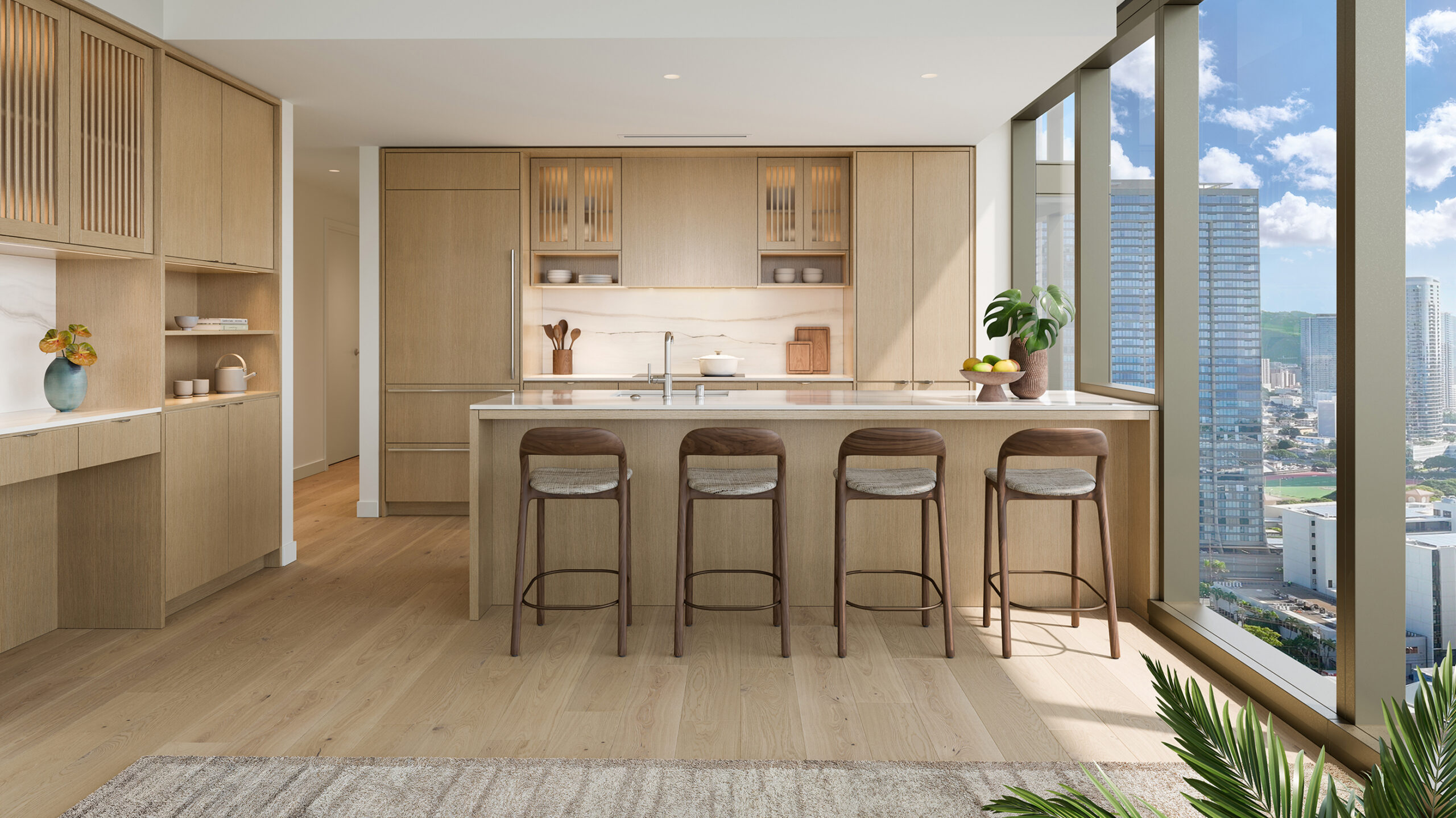 Modern kitchen with light wood cabinets, island with four stools, and large windows showing a cityscape and blue sky. Inspired by Mahana Ward Village residences, this space features a plant and fruit bowl on the counter, with natural light filling the room.