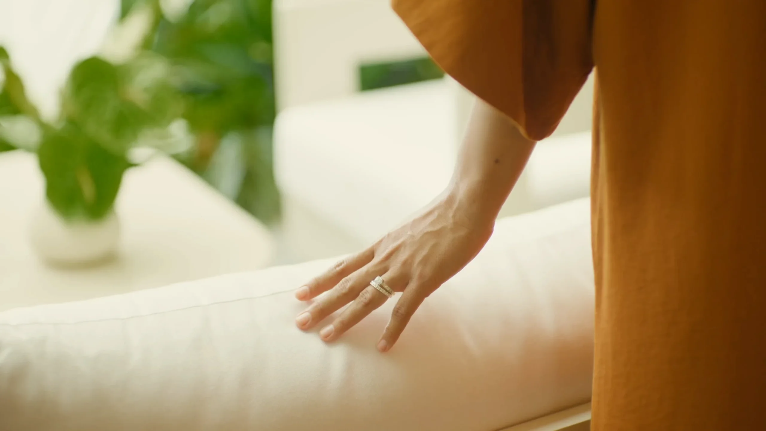 A person wearing an orange sleeve touches a white cushion with their left hand. A silver ring is visible on their ring finger. Green leaves are blurred in the background.